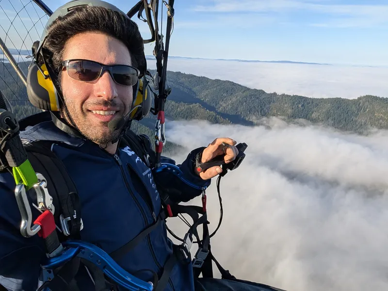 Nimo under his paramotor wing above a bank of fog and forested hills